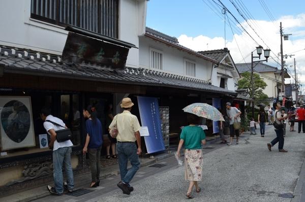 奥には日本家屋が並んでいて、手前の道路を日傘をさした女性や帽子をかぶった男性が歩いている写真